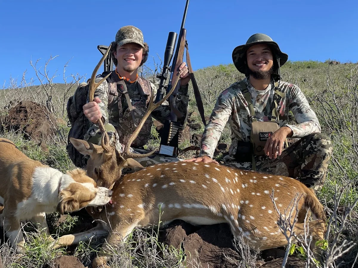 Two hunters kneel behind a harvested axis buck with a dog beside them.