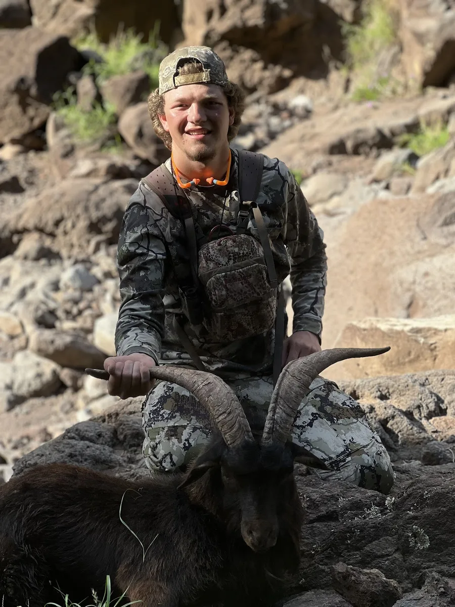 A hunter kneels behind a harvested horned goat on rocky terrain.