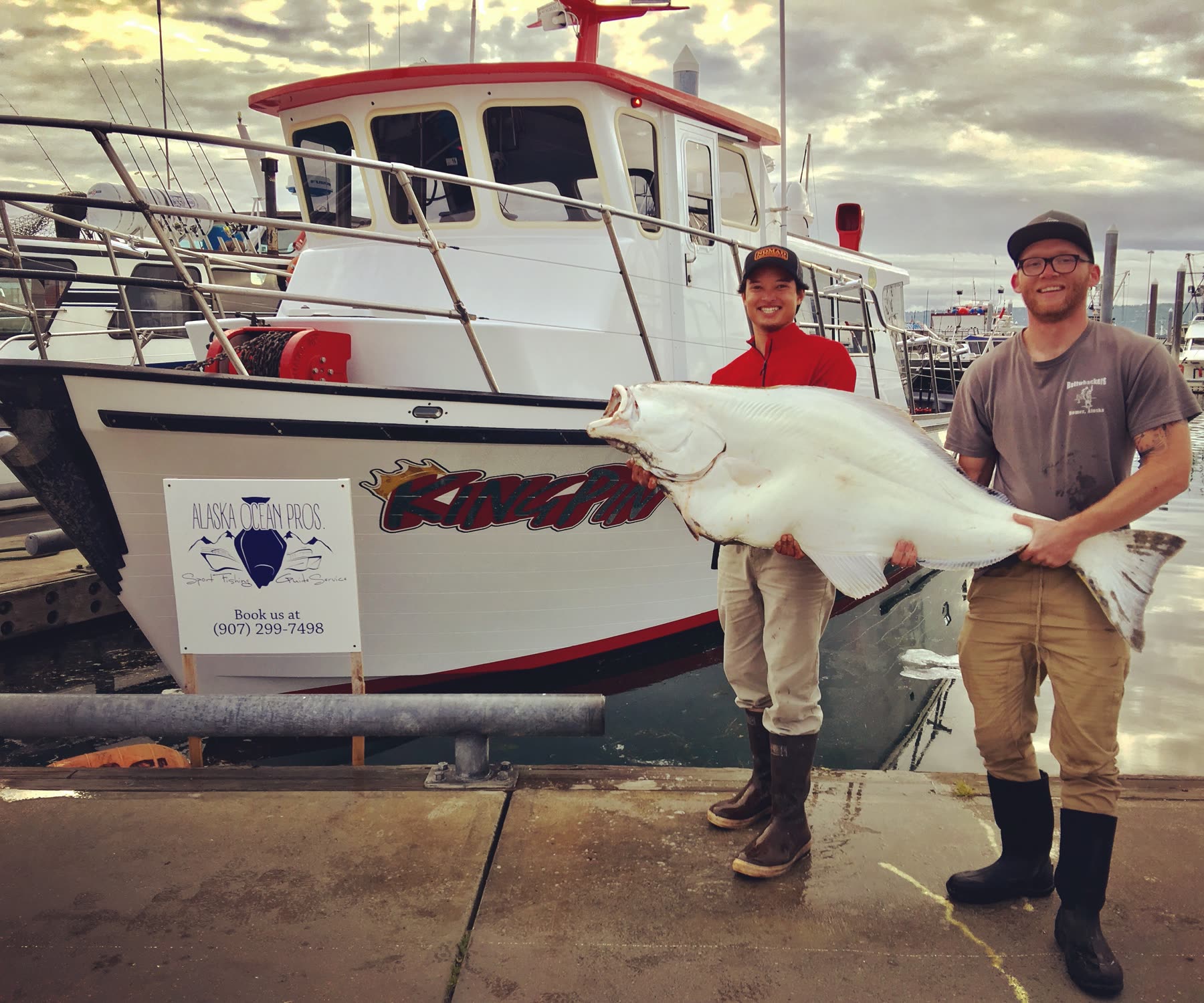 Captain Pono and his first mate stand beside a large halibut and the King Pin boat at an Alaska harbor.